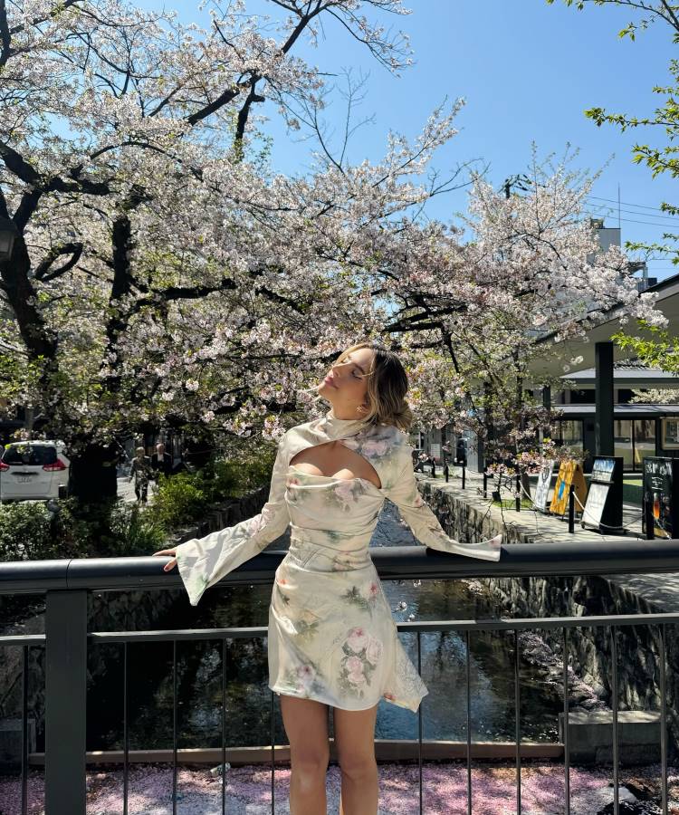 Jovem de cabelos loiros posa em uma ponte cercada por árvores floridas de cerejeira sob um céu azul. Ela usa um vestido floral elegante e olha para cima, transmitindo serenidade em um cenário encantador de primavera.