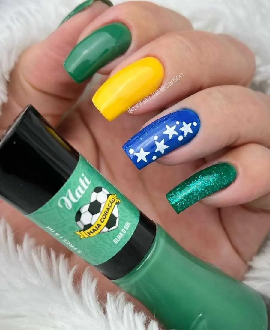 Imagem de mão decoradas com esmalte.  Cores usadas, verde, amarelo, azul e branco. Polegar: verde. Indicador: verde. Dedo Médio, amarelo. Anelar: azul com estrelas brancas e glitter. Mínimo: verde com glitter.