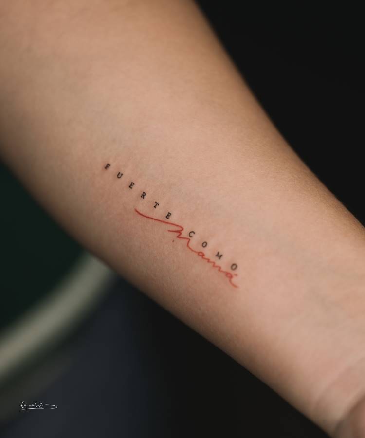 A imagem mostra uma tatuagem no antebraço de uma pessoa. A tatuagem diz "FUERTE COMO" em letras minúsculas pretas, e "Mama" em letras cursivas vermelhas, com o "Ma" sublinhado e as letras se estendendo. A pele é clara e o fundo escuro.








