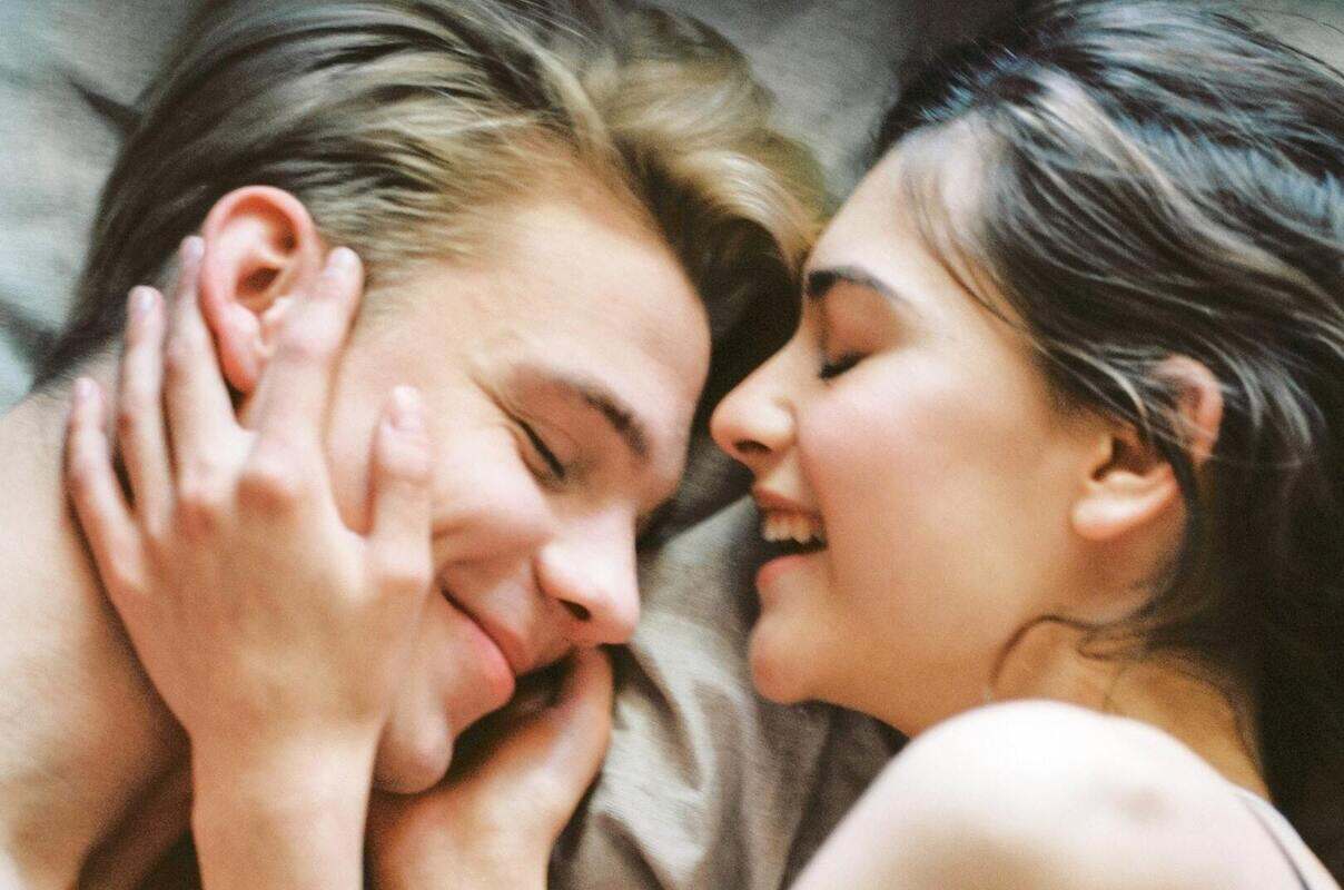 Casal heterossexual formado por homem loiro de pele clara e mulher morena de pele clara sorrindo na cama 