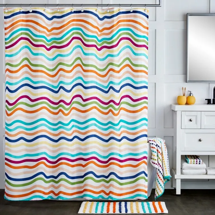 Banheiro iluminado com cortina de chuveiro decorada com listras onduladas multicoloridas, combinando com tapete listrado colorido. Pia branca com espelho retangular, acessórios amarelos e toalhas dobradas. Design alegre e funcional com detalhes organizados.
