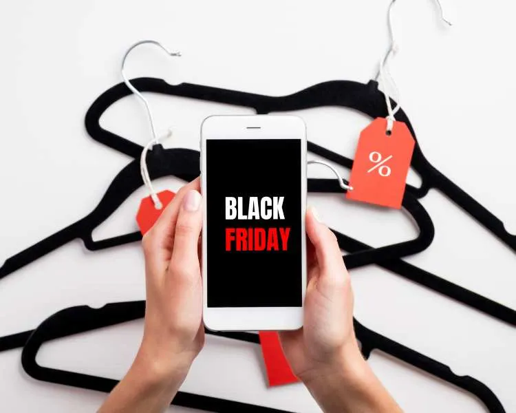 Mão segurando celular com imagem escrito Black Friday de branco e vermelho em fundo preto. No fundo da foto, cabides pretos sobre uma superfície branca