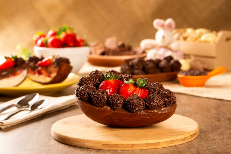 Metade de um ovo de chocolate recheado com morangos frescos e brigadeiros está sobre uma tábua de madeira. Ao fundo, decoração com doces, morangos, trufas e um coelho decorativo compõem um ambiente festivo e acolhedor.