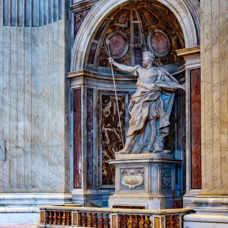 Escultura de mármore em um nicho ornamentado, representando um homem com manto fluido, segurando uma lança. A obra está em um cenário de arquitetura renascentista com colunas, detalhes dourados, e fundo decorado com mármore em tons escuros.