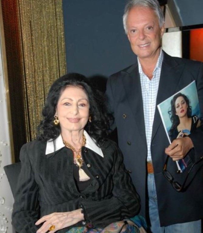 Foto de Carmen Mayrink Veiga e Guilherme Guimarães mais velhos posando lado a lado, ela sentada e ele em pé com um revista Foto de Carmen Mayrink Veiga e Guilherme Guimarães mais velhos posando lado a lado, ela sentada e ele em pé com um revista