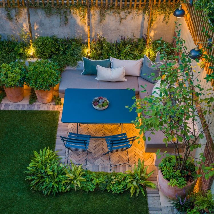 A imagem mostra um jardim moderno e aconchegante, com um sofá em L adornado por almofadas. Uma mesa azul com cadeiras ocupa o centro, cercada por vasos grandes, plantas vibrantes e grama artificial, iluminado por luzes suaves.