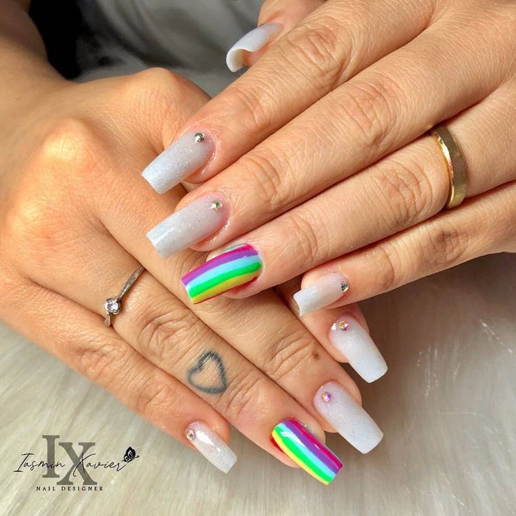 Uma mão com unhas pintadas de diferentes cores. O polegar, indicador e dedo médio possuem esmalte branco com glitter prata e uma pedrinha decorativa próxima à cutícula. O anelar exibe um desenho completo do arco-íris cobrindo toda a unha. Por fim, o dedo mínimo também possui esmalte branco com glitter prata e uma pedrinha decorativa próxima à cutícula.
