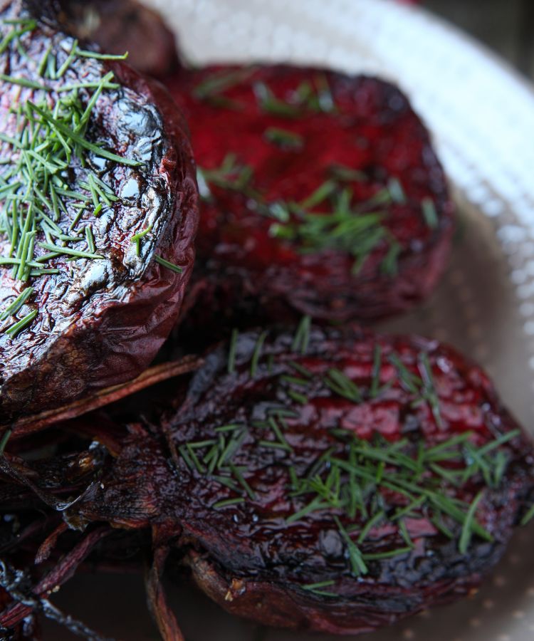 Beterrabas assadas, cortadas ao meio, em um prato claro. As beterrabas têm uma cor roxo-escura e um brilho caramelizado na superfície, finalizadas com cebolinha picada por cima.