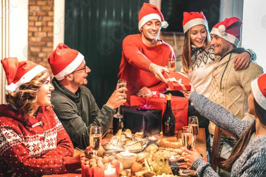 Foto de pessoas de pele clara com gorro de papai Noel, usando suéteres, em volta de mesa com comida e presentes