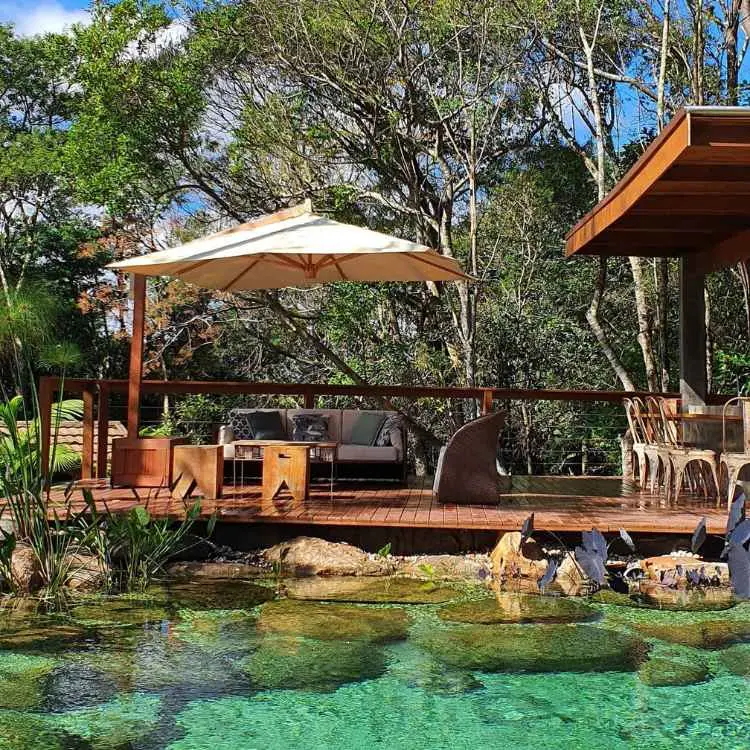 Piscina biológica ao lado de área gourmet, com mesas. cadeiras, sofás e guarda-sol. 