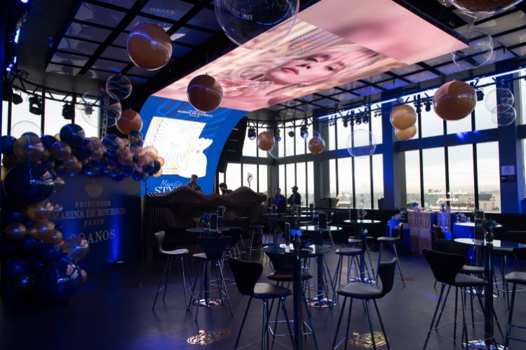 Evento em ambiente moderno e sofisticado, com mesas altas e decoração com balões dourados e azuis. Tela de projeção no teto exibe imagem de modelo. Luzes azuis iluminam o espaço, celebrando "Princesse Marina de Bourbon Paris - 30 anos".