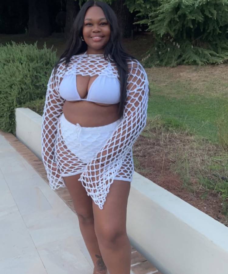 Mulher sorridente com cabelo solto posa usando uma saída de praia de crochê branca. Ela veste um biquíni branco e shorts. Ao fundo, plantas verdes e uma parede de cimento.
