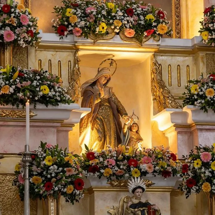 A imagem mostra uma estátua de uma figura feminina com um manto, provavelmente a Virgem Maria, segurando uma criança, possivelmente o menino Jesus. Eles estão cercados por flores coloridas e situados em um altar decorado com elementos dourados e velas acesas, criando um ambiente sagrado e reverente.

