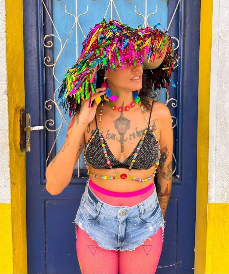Foto de mulher usando look confortável de Carnaval: meia calça rosa, micro short jeans, top de miçangas e chapéu de franjas