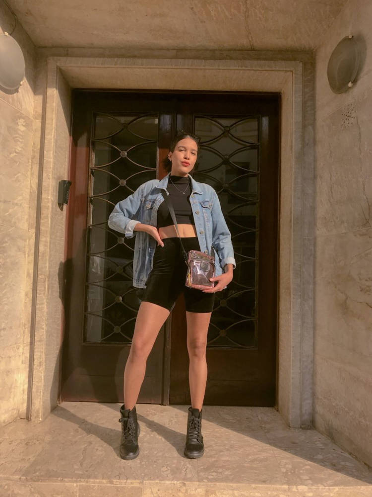 Mulher de pele morena usando cropped preto, jaqueta jeans, bermuda ciclista preta, shoulder bag rosa e coturno preto 