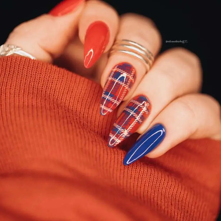 Mão com unhas longas no formato amendoado. Duas unhas são vermelhas sólidas, duas têm um padrão xadrez vermelho e azul, e uma é azul sólida. A mão está sobre um tecido vermelho.