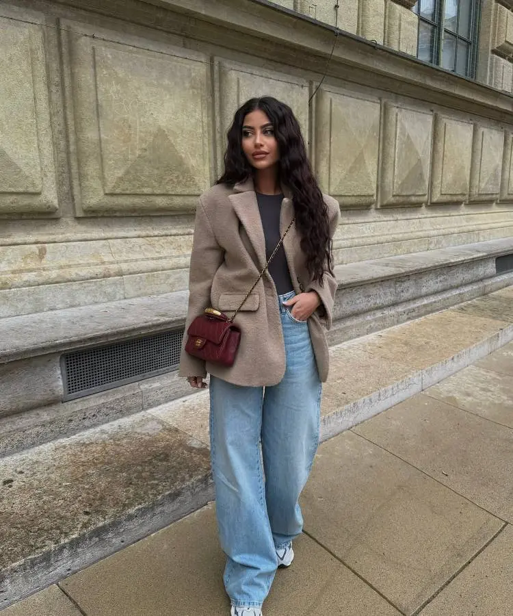 Uma mulher com cabelo longo e ondulado veste um blazer bege oversized sobre uma camiseta cinza, combinando com jeans largos de lavagem clara e tênis brancos. Ela carrega uma pequena bolsa de ombro vermelha escura. O fundo é uma parede de pedra com janelas.