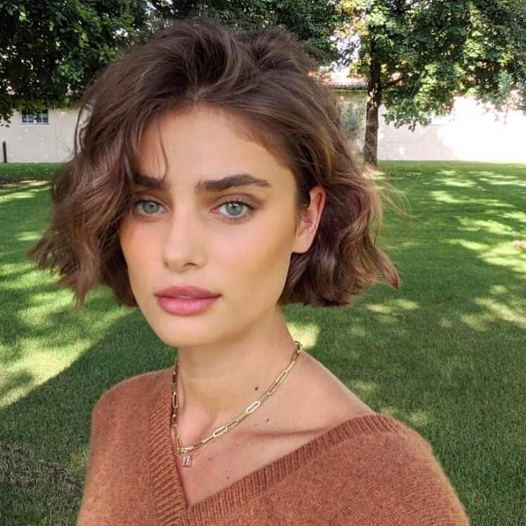 Mulher com cabelo curto estilo bob ondulado, castanho, com volume natural. Pele clara, olhos verdes realçados, sobrancelhas grossas e lábios rosados. Usa suéter marrom, colar dourado, e está ao ar livre em um gramado ensolarado com árvores ao fundo.