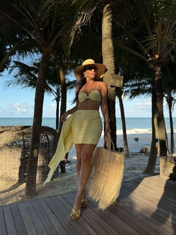 Mulher de pele clara na praia usando biquini e saia amarelos, além de chapéu e bolsa de palha com franjas