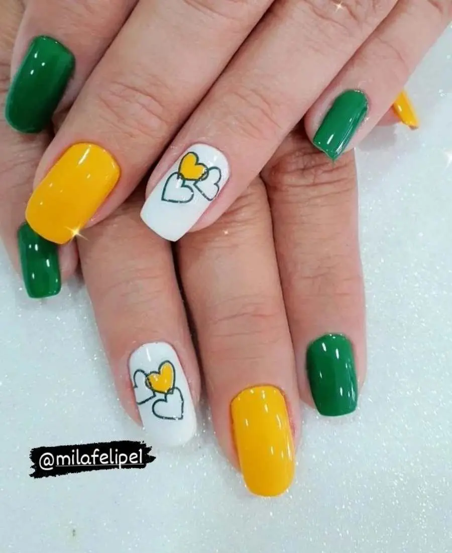 Imagem de mão com unhas decoradas para a Copa do Mundo 2022. Cores usadas, verde, amarelo e branco. Polegar: amarelo. Indicador: verde. Dedo Médio, amarelo. Anelar: branco com adesivo de corações, um sendo amarelo. Mínimo: verde.