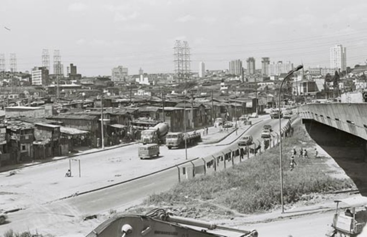 A imagem mostra uma área periférica de São Paulo nos anos 1960, com habitações precárias contrastando com prédios ao fundo. Um viaduto em construção destaca o crescimento urbano, enquanto ônibus e pedestres ocupam ruas simples em um cenário desigual.
