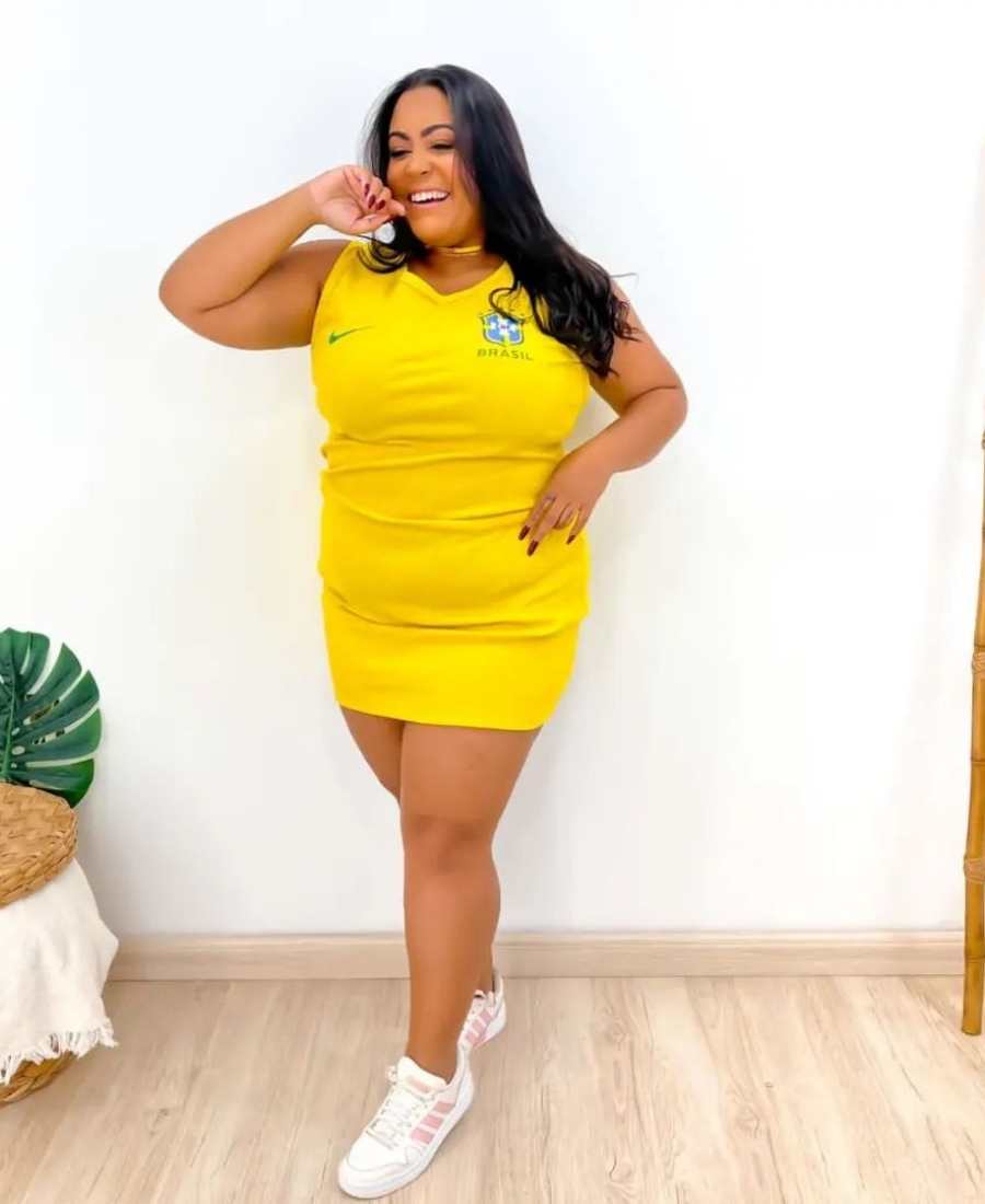 Imagem com fundo de parede e plantas . No centro, mulher usando roupa copa do mundo: vestido amarelo do Brasil + tênis branco e rosa.