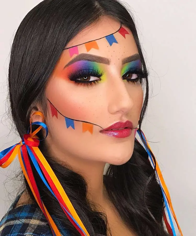 Uma mulher com maquiagem marcante para Festa Junina, com sombras de olhos em tons de arco-íris e sardas falsas. Desenhos de bandeirinhas coloridas adornam sua testa e bochechas, e seu cabelo tem tranças e fitas coloridas.
