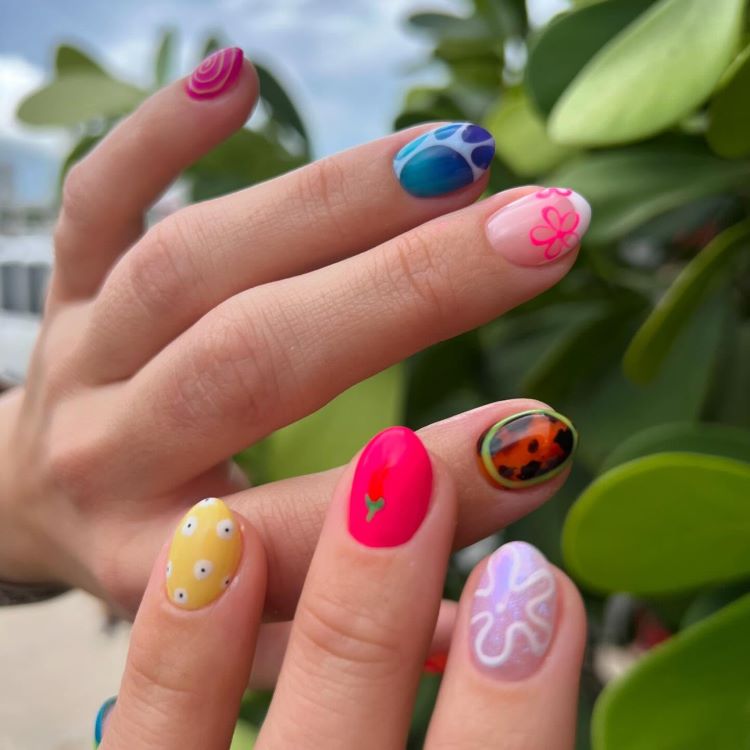 Unhas decoradas com designs variados e coloridos: espiral rosa, gotas azuis, flor rosa, joaninha, coração vermelho, padrão abstrato e margaridas em amarelo. Fundo natural com folhas verdes desfocadas, criando contraste vibrante e alegre para os detalhes.