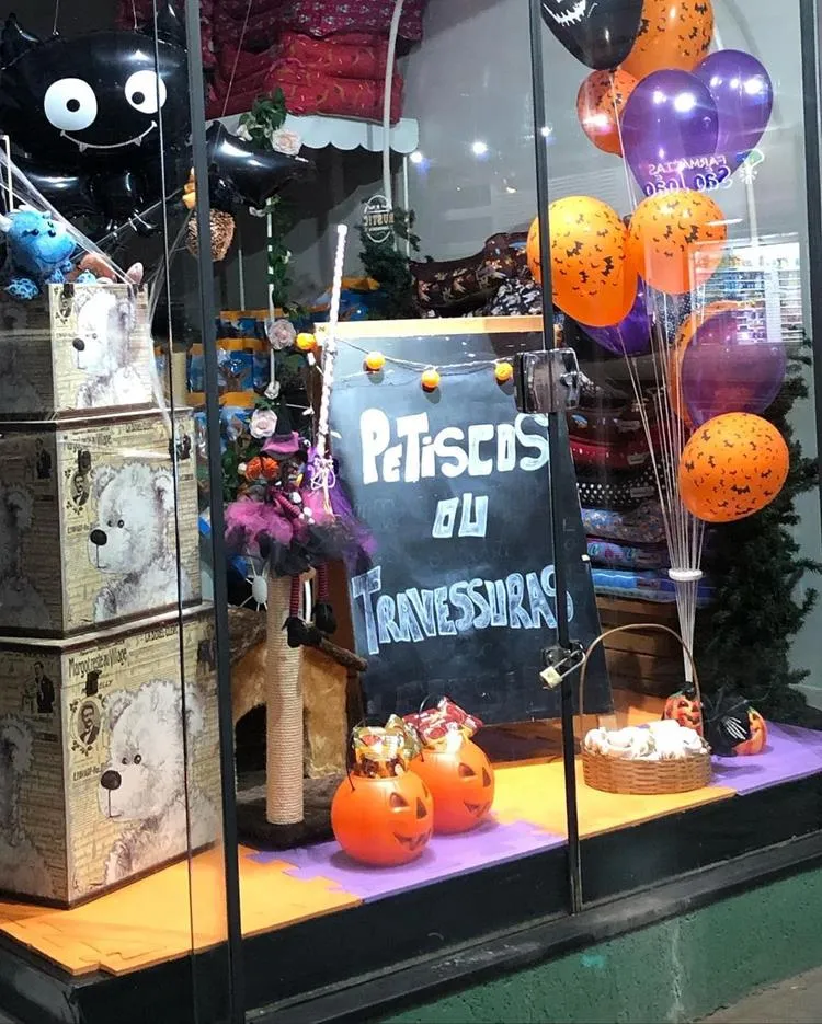 Vitrine de pet shop com bexigas de halloween, caixas com desenho de urso, potes em formato de abóbora e lousa com frase "Petiscos ou travessuras"