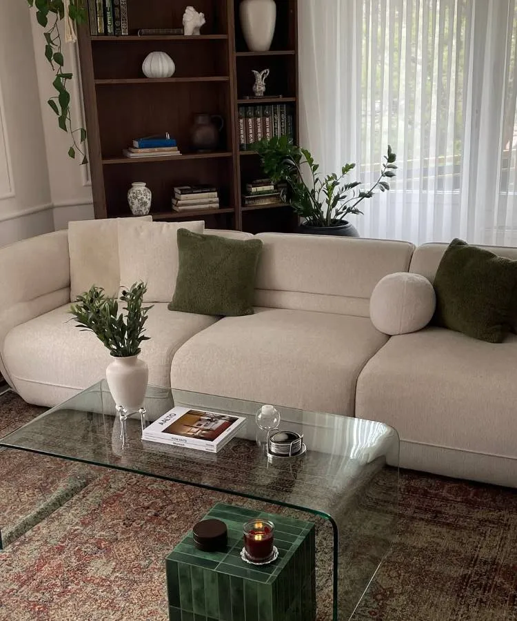 Uma sala de estar elegante com sofá bege, almofadas para sala verdes e redondas. Uma mesa de centro de vidro curvado exibe um vaso com folhagens e livros. Ao fundo, uma estante escura com livros e objetos decorativos. Cortinas brancas e plantas penduradas completam o ambiente.







