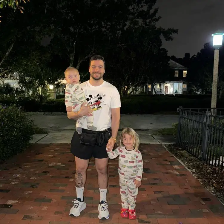 A imagem noturna mostra um homem sorridente de camiseta do Mickey, segurando um bebê no colo e de mãos dadas com uma criança pequena, ambos de pijama listrado. Eles estão em um caminho de tijolos, com casas e árvores iluminadas ao fundo.







