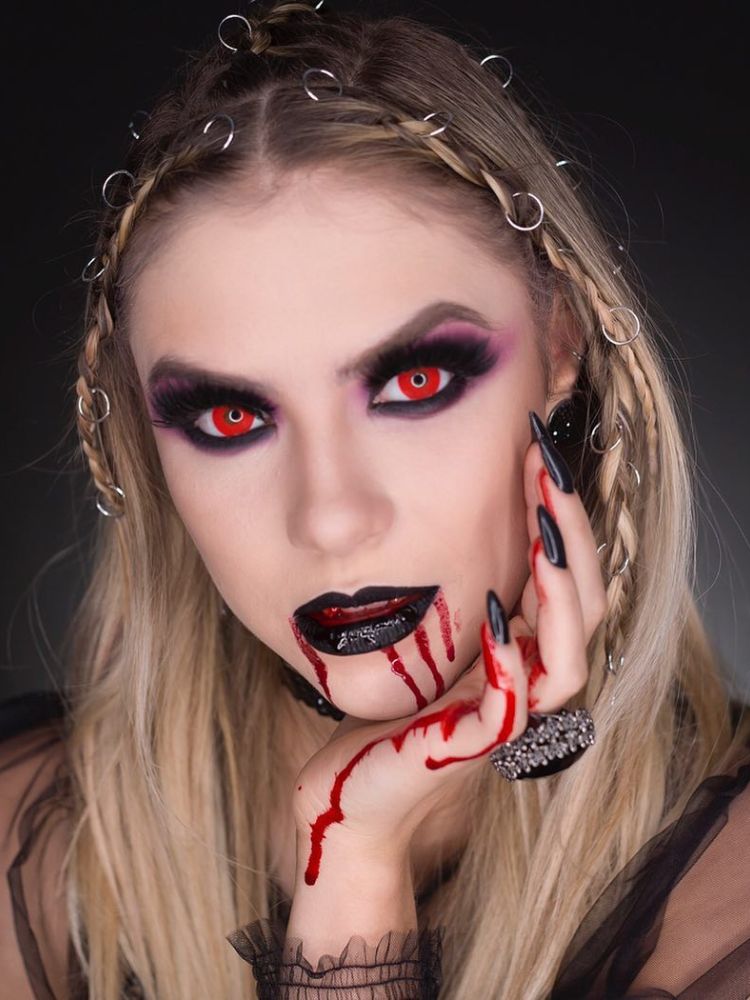 Mulher de pele clara com lente vermelha no olho, batom escuro e boca escorrendo sangue artificial