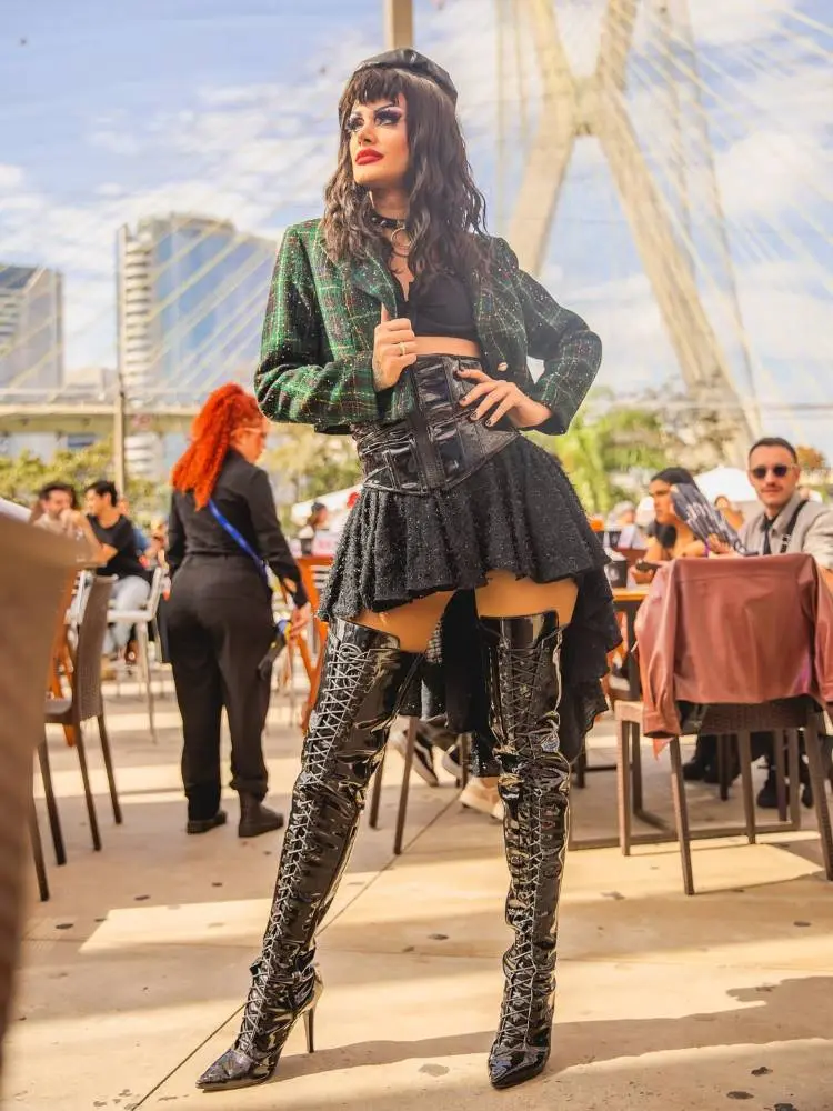 Drag Queen posa ao ar livre em frente a ponte estaiada, cercada por pessoas em mesas. Usa saia preta brilhante, top e jaqueta verde xadrez. Botas pretas altíssimas de verniz chamam atenção. Cabelo escuro com franja e maquiagem marcante completam look.