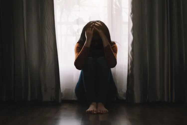 A imagem mostra uma mulher sentada no chão, de frente para uma janela com cortinas. Ela está em silhueta contra a luz, com as mãos no rosto, sugerindo angústia ou choro. O ambiente parece íntimo, com foco na figura e nas cortinas que a emolduram.
