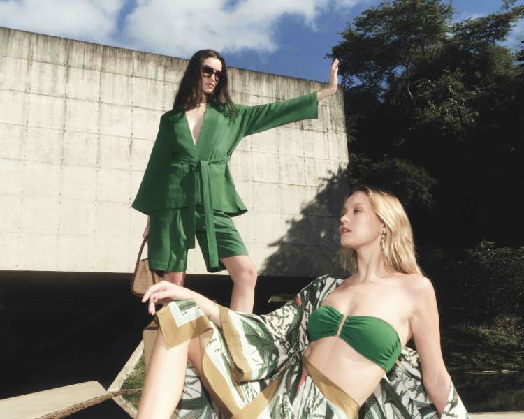 Duas modelos em poses descontraídas. Uma, sentada, veste um top de biquíni verde e shorts estampados. A outra, em pé, usa um conjunto de shorts e blazer verdes, com óculos de sol.Coleção Lenny Niemeyer + Inhotim.