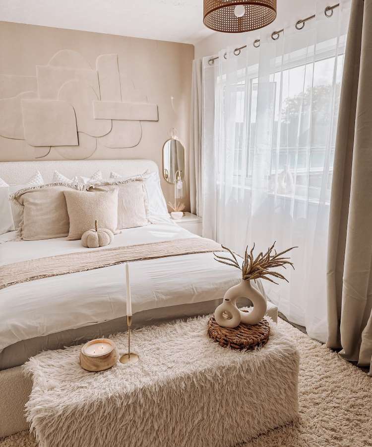 Quarto de casal com espelho oval pequeno na parede sob mesinha de canto. Cores predominantes: off-white, dourado, palha