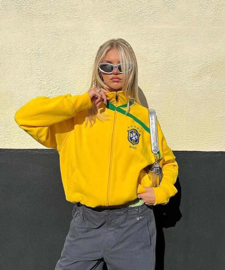 Imagem com fundo de parede preto e branco. No centro, mulher usando blusa de frio amarela do Brasil + calça + bolsa prata