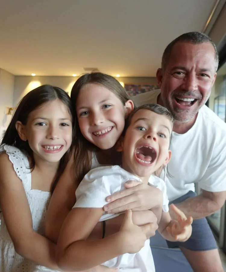 Um pai sorridente e de camiseta branca posa com seus três filhos. Duas meninas sorriem e abraçam o irmão mais novo, que faz uma careta divertida. Nomes diferentes de filhos de famosos: a cena captura a alegria e a espontaneidade de uma família unida.

