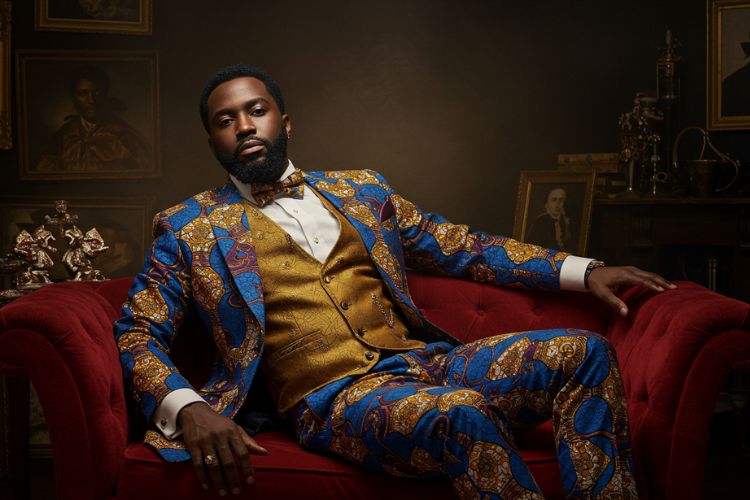 Homem negro sentado em um sofá vermelho veste um elegante terno de estampa africana azul e dourada, combinado com colete dourado e gravata borboleta. Ao fundo, quadros e objetos decorativos em ambiente sofisticado.