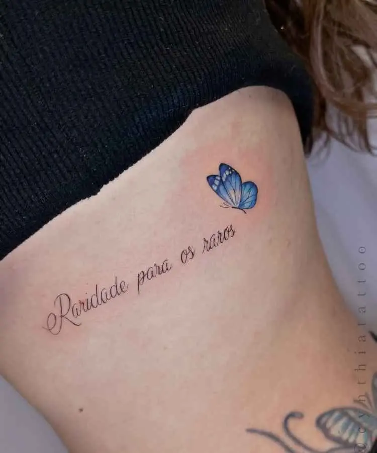 A imagem mostra uma tatuagem delicada na lateral do corpo com a frase cursiva "Raridade para os raros". Ao lado, há uma borboleta azul detalhada e vibrante. A pele está levemente avermelhada, evidenciando um trabalho recente e bem-feito.