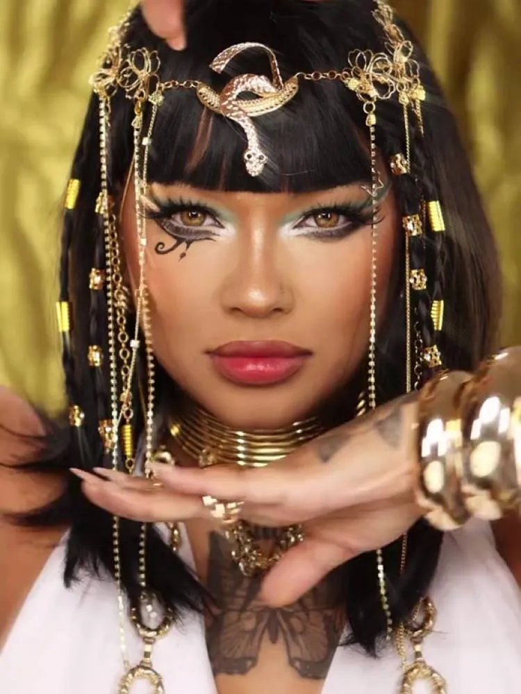Uma mulher com maquiagem elaborada e acessórios inspirados em Cleópatra makeup trend. Ela usa uma peruca preta com tranças e adereços dourados, incluindo uma tiara de cobra e correntes penduradas. A maquiagem dos olhos é dramática, com delineado gráfico e um "Olho de Hórus" no rosto.

