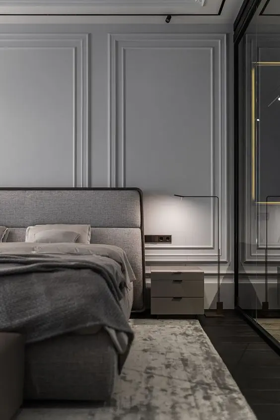 Quarto de casal com piso escuro, cama cinza, armário espelhado, mesa de cabeceira cinza e parede bege com três tamanhos de moldura 