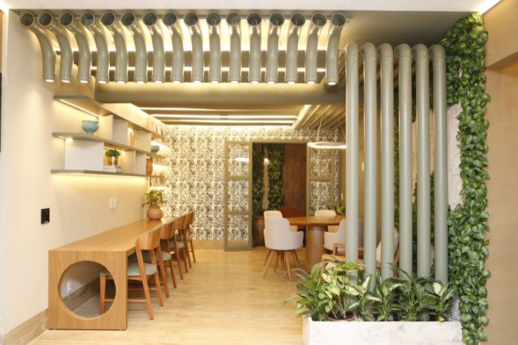 Espaço de coworking com cadeiras e mesas de madeira, poltrona, piso de madeira e canos de PVC cinza como luminárias
