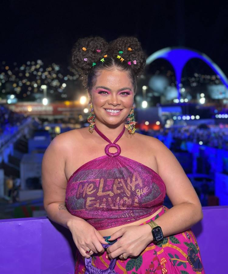 A mulher sorri com maquiagem vibrante rosa e penteado com coques decorados por mini acessórios coloridos. Usa um top roxo com a frase "Me Leva que Eu Vou", brincos chamativos e está em um cenário carnavalesco iluminado.
