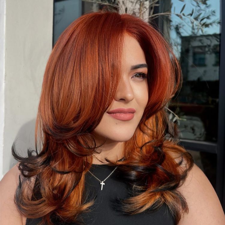 Mulher com cabelos longos em tons de ruivo acobreado mesclado com pontas escuras, finalizados com ondas sutis. Ela usa maquiagem suave, batom nude e veste blusa preta. A luz natural destaca o brilho dos fios em ambiente externo.