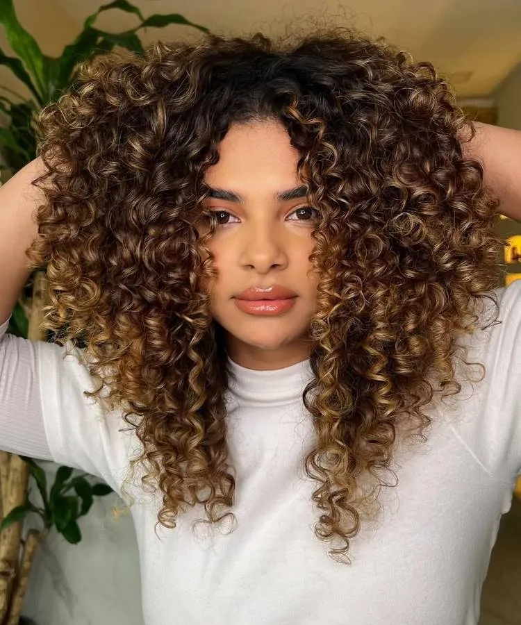 A mulher com cabelos encaracolados castanhos mel e luzes loiras. Ela está olhando diretamente para a câmera com as mãos atrás da cabeça, vestindo uma blusa branca.