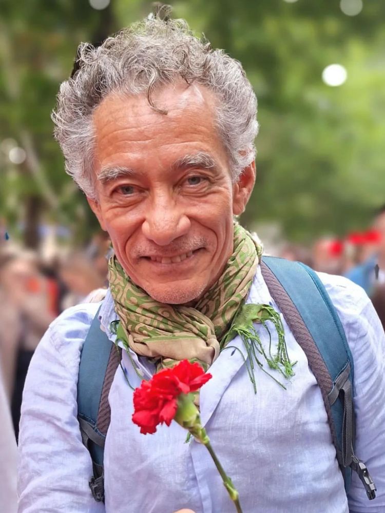 Foto de Chico Diz usando camisa social clara, cachecol verde, segurando flor vermelha
