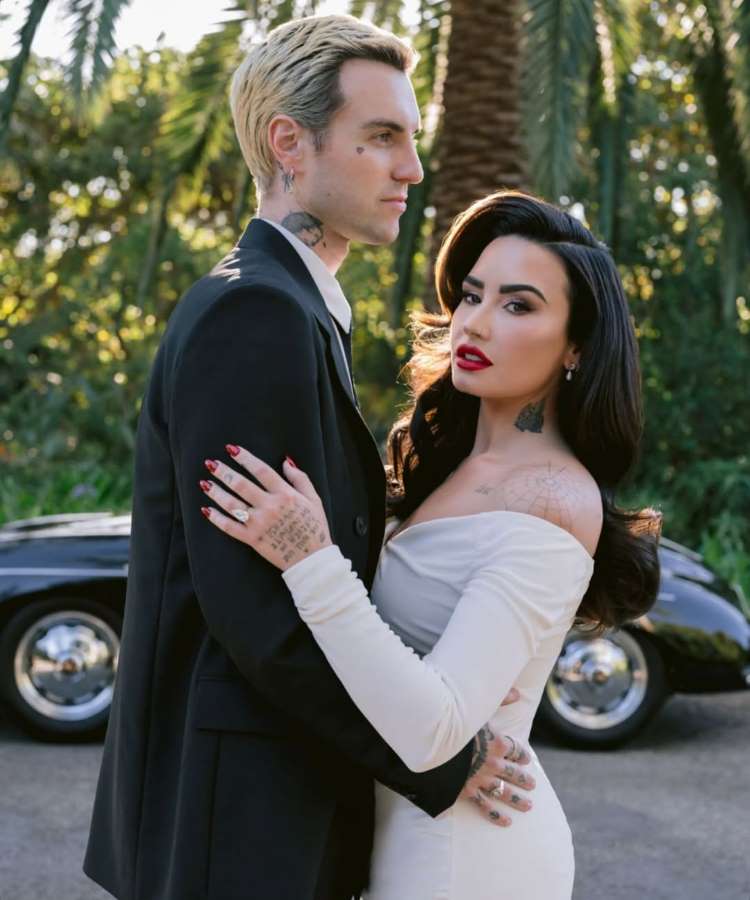 Demi Lovato e Jordan Lutes estão de pé em um cenário exterior. Jordan, com cabelo loiro, veste um terno preto e olha para o lado. Demi, de cabelo escuro e vestido branco de ombros caídos, olha para a câmera enquanto o abraça. Um carro escuro está ao fundo.