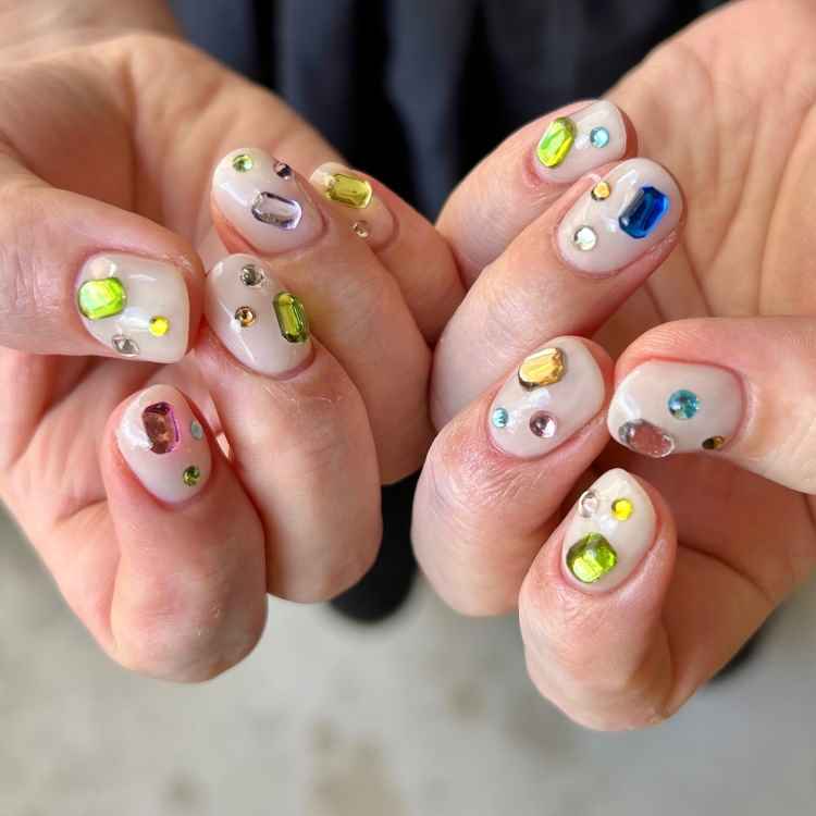 Unhas curtas com esmalte nude, decoradas com pedras coloridas em vários formatos e cores, como verde, azul e rosa. As mãos estão levemente fechadas, mostrando o design único e criativo. O fundo é neutro, destacando o brilho das gemas.