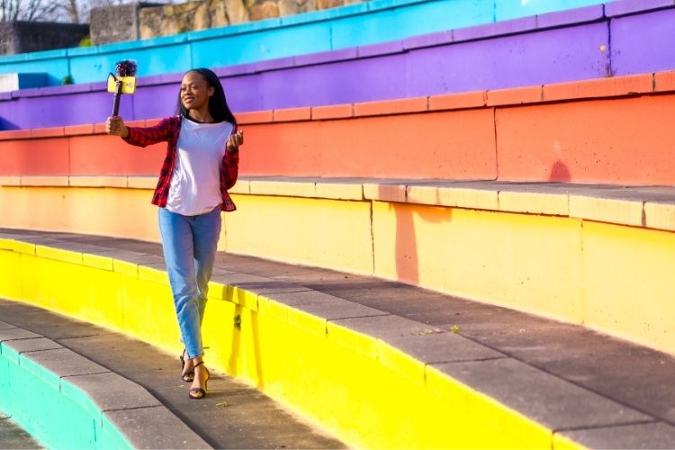 Uma mulher sorridente, de camisa xadrez vermelha e jeans, caminha num anfiteatro colorido com degraus em tons de amarelo, laranja, roxo e azul, segurando um dispositivo de gravação.







